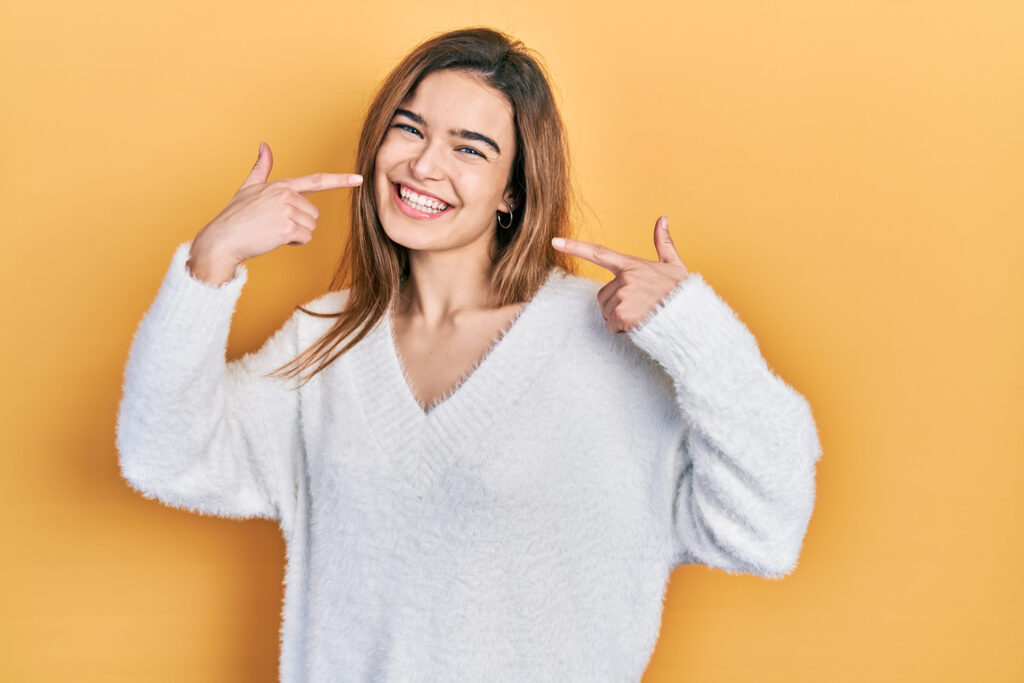 A smiling young woman with brown hair, wearing a white fuzzy V-neck sweater, points to her wide, happy smile with both index fingers. The background is a solid yellow/orange color.
