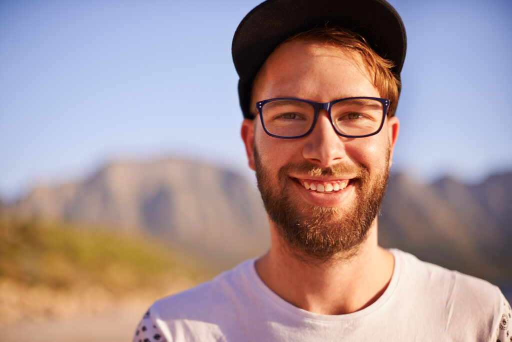 A bearded man wearing glasses and a cap smiles outdoors with mountains and a blue sky in the background.