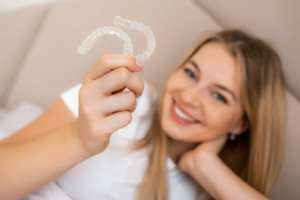A close-up shot focusing on a woman's hand holding a clear dental aligner, with her smiling face blurred in the background.